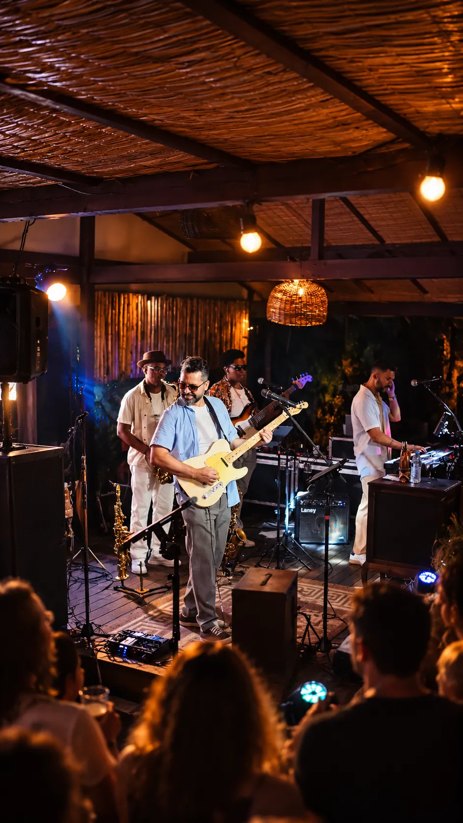 Pablo Curty tocando guitarra e sorrindo no palco, com a banda atrás e o público em primeiro plano.